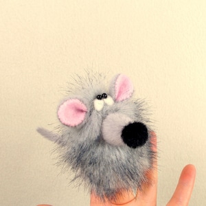 Peut inclure: Une marionnette de doigt en forme de souris, grise et blanche, avec des oreilles roses et des yeux noirs. La souris a un nez noir et une queue noire.
