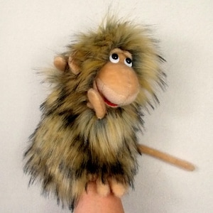 Può includere: Un burattino a forma di scimmia. Il burattino ha una faccia marrone, grandi occhi neri e una bocca rossa. La sua pelliccia è un misto di marrone e nero. Il burattino è progettato per essere infilato sulla mano per giocare.