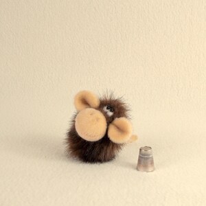 Puede incluir: Un animal de peluche marr&oacute;n y beige con orejas grandes y una nariz peque&ntilde;a y redonda. El juguete est&aacute; sentado sobre una superficie blanca junto a un dedal de plata.