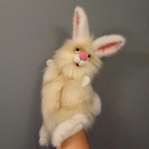 May include: A plush rabbit hand puppet with long, cream-coloured faux fur, tall ears with pink inner lining, and a bright pink nose. The puppet features dark, round eyes, a white muzzle, and a small open mouth, designed for use as a hand-operated toy.