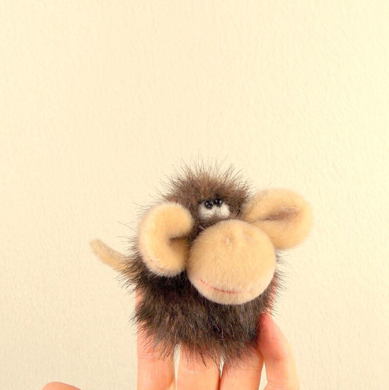 Puede incluir: Un peque&ntilde;o mono de peluche marr&oacute;n y beige con orejas grandes. El mono est&aacute; sentado en una mano.