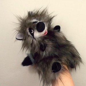 Pode incluir: Uma marioneta de mão de guaxinim de peluche com pelo sintético comprido e desgrenhado em tons de cinzento e preto. A marioneta tem uma máscara facial branca com manchas pretas nos olhos, um nariz preto redondo e pequenas patas pretas.