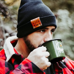 May include: A man wearing a black beanie with a leather patch that says "NW" is drinking from a green enamel mug with the text "Escape to the Wild Est. 1987". He is wearing a red and black plaid flannel shirt.