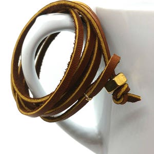 May include: A brown leather wrap bracelet with a gold metal clasp. The bracelet is wrapped around a white ceramic mug.