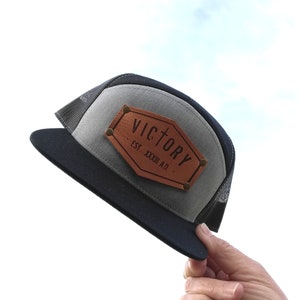 May include: A gray and black trucker hat with a brown leather patch that says "VICTORY EST. XXXIII A.D."