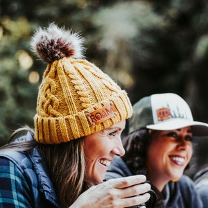 May include: Two women are smiling and holding mugs. One woman is wearing a yellow knit beanie with a brown leather patch that says "ROAM". The other woman is wearing a green and white baseball cap with a brown leather patch.