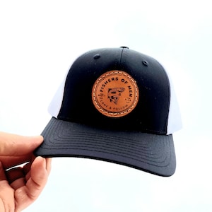 May include: Black and white trucker hat with a brown leather patch that says "Fishers of Men" with a fish illustration. The text on the patch says "Come & Follow".