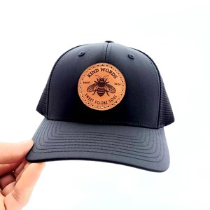 May include: Black trucker hat with a brown leather patch featuring a bee and the text "Kind Words Prov. 16:24 Sweet to the Soul".