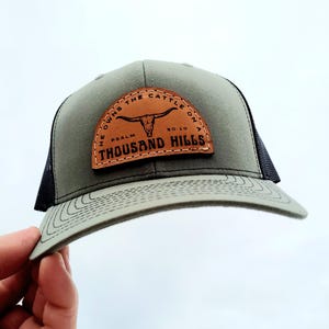 May include: A green and black trucker hat with a brown leather patch that says "He owns the cattle on a thousand hills, Psalm 50:10". The patch features a stylized bull's head.