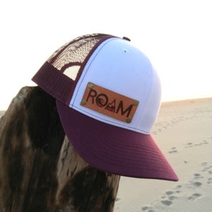 May include: A white and burgundy trucker hat with a brown leather patch that says "ROAM" in white lettering. The patch has a mountain graphic.