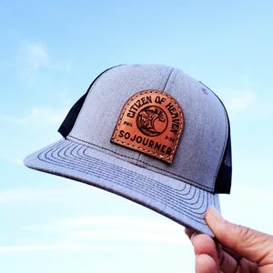 May include: Gray trucker hat with a black mesh back and a curved brim. A brown leather patch on the front displays the text "Citizen of Heaven" and "Sojourner" with a graphic. The hat is held up against a blue sky.