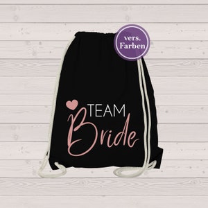 Może przedstawiać: Czarny plecak ze sznurkiem z napisem "TEAM Bride" w różowym skrypcie i różowym sercem. Torba ma białe sznurki i okrągłą fioletową naklejkę w prawym górnym rogu z napisem "vers. Farben".