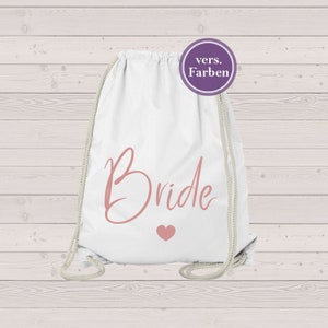 Könnte beinhalten: Weißer Kordelzugbeutel mit dem Wort "Bride" in rosa Schrift und einem kleinen rosa Herz. Die Tasche hat einen Kordelzugverschluss und einen runden lila Aufkleber mit deutschem Text.