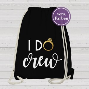 Peut inclure: Sac de sport noir avec l'inscription "I DO crew" en écriture blanche et un motif d'anneau doré. Le sac a des cordons de serrage blancs et un cercle violet avec du texte allemand.
