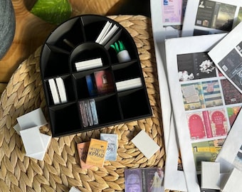 Könnte beinhalten: Ein schwarzer, bogenförmiger Organizer mit mehreren Fächern, der kleine Bücher und weiße Notizkarten enthält. Miniaturbücher mit farbenfrohen Einbänden und russischem Text sind verstreut, zusammen mit bedruckten Blättern mit Buchumschlägen. Eine kleine, weiße Topfpflanze sorgt für einen Hauch von Grün.