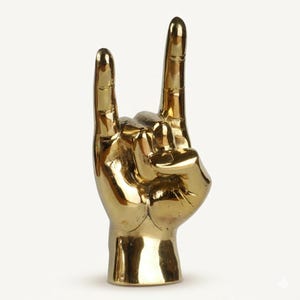 May include: A polished gold-colored metal sculpture shaped like a human hand making the rock and roll sign. The hand features an upright index finger and pinky finger, with the middle and ring fingers folded down toward the palm, showing smooth surfaces.