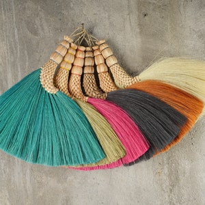 May include: A set of seven colorful brooms made of natural fibers, with the handles tied together. The brooms are arranged in a fan shape, with the colors ranging from teal to beige to orange to cream.