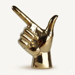 May include: A polished brass sculpture shaped like a human hand with the index finger extended upward and the thumb pointing outward. The metallic surface is reflective and smooth, capturing light across the fingers and the cylindrical base.
