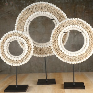 Op de afbeelding: Drie witte schelpenkransen in verschillende maten op zwarte metalen standaards. De kransen zijn gemaakt van kleine, witte schelpen en zijn in een cirkel gerangschikt.