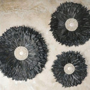 Puede incluir: Tres piezas de decoración de pared de plumas negras con un centro de concha blanca. Las piezas están dispuestas en un patrón escalonado sobre un fondo marrón claro.