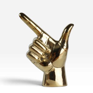 Op de afbeelding: Een sculptuur van gepolijst goudkleurig metaal in de vorm van een hand met de wijsvinger uitgestrekt en de duim omhoog gericht. Het oppervlak is glad en reflecterend en vangt het licht op de vingers en de basis van de polsstructuur.