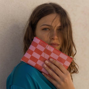 Peut inclure: Une femme tient une pochette zippée à carreaux rose et rouge devant son visage. La pochette a une fermeture éclair argentée.