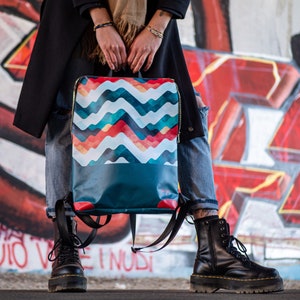 Può includere: Uno zaino blu e bianco con motivo e dettagli in rosso, giallo e arancione. Lo zaino ha una chiusura con cerniera e spallacci regolabili.