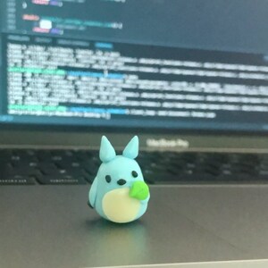 May include: A light blue figurine of a cartoon character with large ears, black eyes, and a cream-colored belly. The figurine is holding a small green object. The background is blurred, showing a laptop and a screen with code.