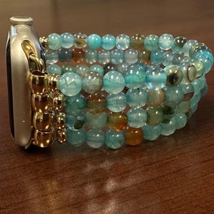 May include: A silver smartwatch with a gold-coloured attachment is connected to a multi-strand bracelet. The bracelet is made of round, translucent beads in shades of blue, green, and brown. The beads are strung together to create a layered effect.