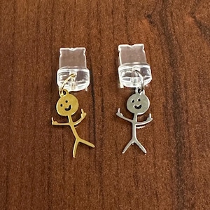 Puede incluir: Dos colgantes de amuleto, uno dorado y otro plateado, cada uno con una figura de palo con una cara sonriente y los dedos medios levantados. Los amuletos están unidos a soportes de plástico transparente.