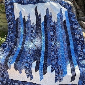 May include: A blue and white quilt with a geometric pattern of vertical stripes and angled lines. The quilt is made of fabric with a celestial print featuring stars, moons, and other celestial bodies.
