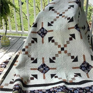 May include: A white quilt with a brown and black geometric pattern. The quilt has a white background with brown and black squares and triangles. The quilt is edged with a brown and black border.