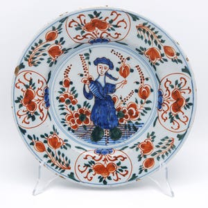 May include: A round ceramic plate featuring a central hand-painted illustration of a figure in blue clothing surrounded by orange and green floral motifs. The rim is decorated with repeating orange and green botanical patterns on a white base.