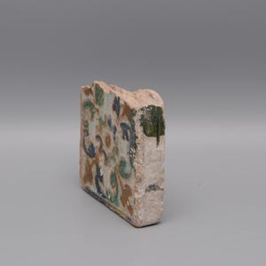 May include: A fragment of a decorative tile with a floral design in shades of blue, green, and brown. The tile has a rough, aged appearance, with visible wear and tear. The edges are uneven, suggesting it is a piece of a larger tile.