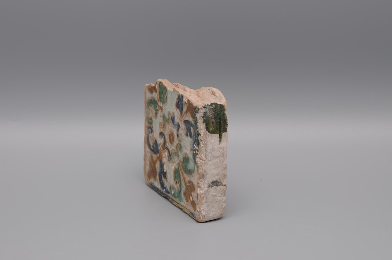 May include: A fragment of a decorative tile with a floral design in shades of blue, green, and brown. The tile has a rough, aged appearance, with visible wear and tear. The edges are uneven, suggesting it is a piece of a larger tile.