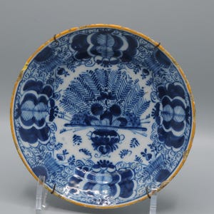 May include: Antique blue and white ceramic plate with a floral design. The plate features a central motif of a stylized floral arrangement and a decorative border. The rim has a gold-colored edge. The plate is displayed on a clear stand.