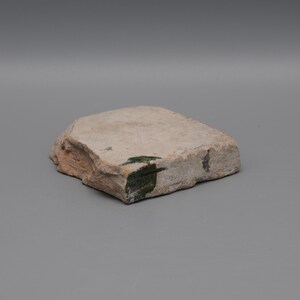 May include: A roughly rectangular, light-colored stone with a textured surface. The stone has a natural, weathered appearance with some areas of green and brown coloration. The edges are uneven, giving it a raw, organic look.