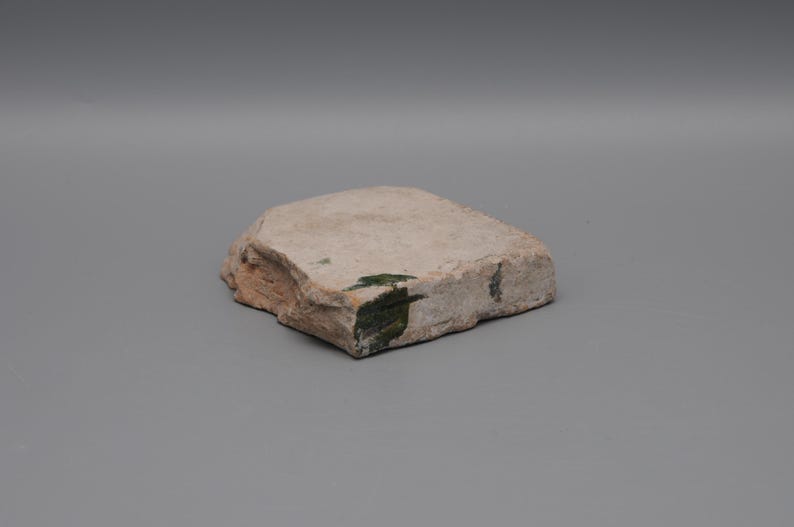 May include: A roughly rectangular, light-colored stone with a textured surface. The stone has a natural, weathered appearance with some areas of green and brown coloration. The edges are uneven, giving it a raw, organic look.