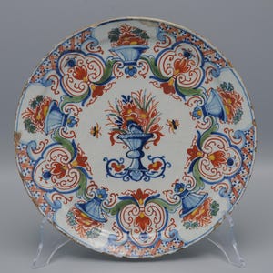 May include: A white ceramic plate with a blue and red floral design. The plate has a raised rim and a scalloped edge.