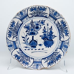 Op de afbeelding: Een ronde keramische bord met een witte basis en gedetailleerde, met de hand geschilderde blauwe ontwerpen. Het midden toont twee vazen gevuld met bloemstukken, terwijl de rand is versierd met herhalende patronen van bladeren en fruit.