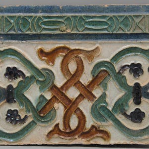 May include: A ceramic tile with a white background and a brown, green, and black geometric design. The design features a central knot with intricate floral patterns.