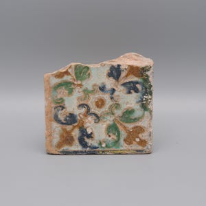 May include: A fragment of a decorative tile with a floral pattern. The tile features a light blue background with green, blue, and brown floral designs. The edges are rough and show the original clay.