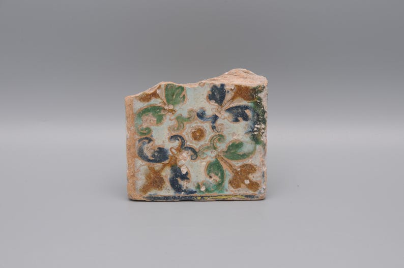 May include: A fragment of a decorative tile with a floral pattern. The tile features a light blue background with green, blue, and brown floral designs. The edges are rough and show the original clay.