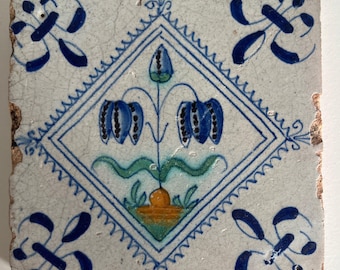 May include: A white ceramic tile with a blue and white diamond pattern. Inside the diamond is a stylized flower with blue, green, and orange paint.