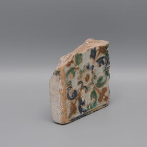 May include: A fragment of a decorative tile with a floral pattern in shades of blue, green, and brown. The tile has a rough, aged appearance, suggesting it is an antique or vintage piece. The background is a solid gray.