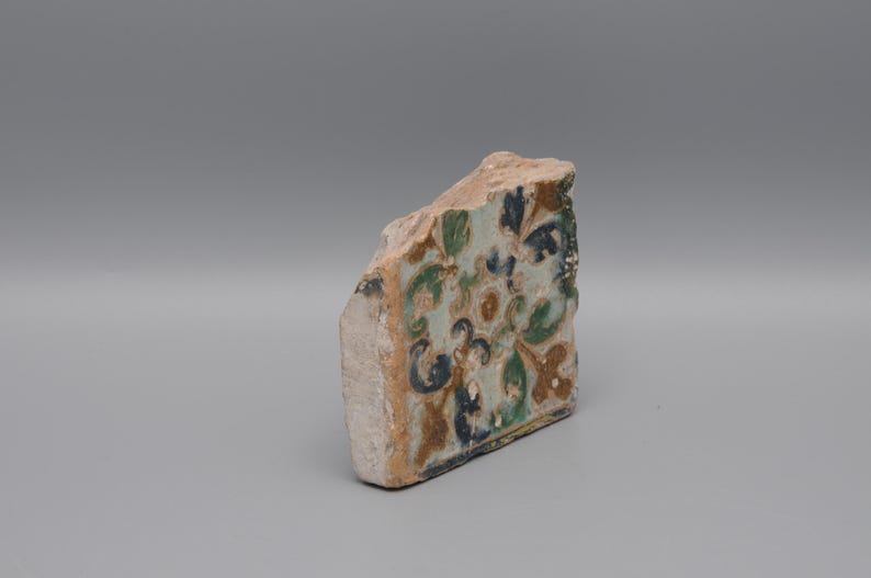 May include: A fragment of a decorative tile with a floral pattern in shades of blue, green, and brown. The tile has a rough, aged appearance, suggesting it is an antique or vintage piece. The background is a solid gray.