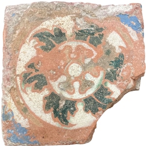 Op de afbeelding: Een fragment van een keramische tegel met een cirkelvormig ontwerp. Het ontwerp heeft een wit centrum met een groen en wit bloemmotief eromheen. De tegel is gebroken en heeft een roodbruine kleur.