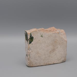 May include: A rectangular, rough-hewn stone block with a light beige color and a textured surface. Patches of green are visible on one side. The stone has an irregular, natural shape, suggesting it is a decorative or craft element.