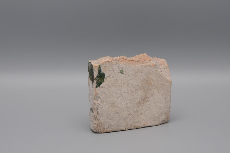 May include: A rectangular, rough-hewn stone block with a light beige color and a textured surface. Patches of green are visible on one side. The stone has an irregular, natural shape, suggesting it is a decorative or craft element.