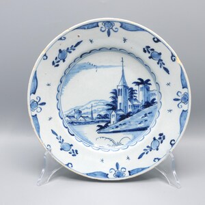 May include: A round ceramic plate featuring a white base with blue hand-painted illustrations. The center depicts a landscape with a church, trees, and hills. The rim is decorated with a repeating blue floral pattern and small star-like accents.